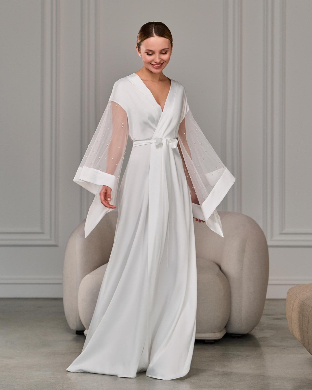 Bride in long white satin robe with pearl-detailed sheer sleeves standing gracefully