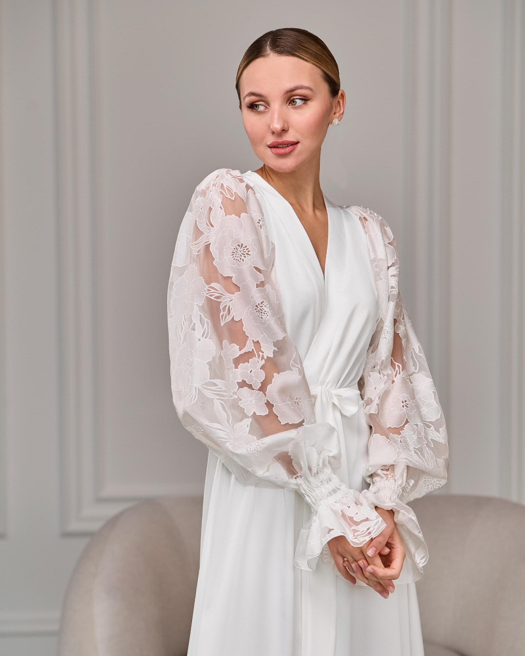 Bride sitting on a beige couch, smiling and gently touching her face while wearing the long ivory robe with sheer floral puff sleeves and a front slit.
