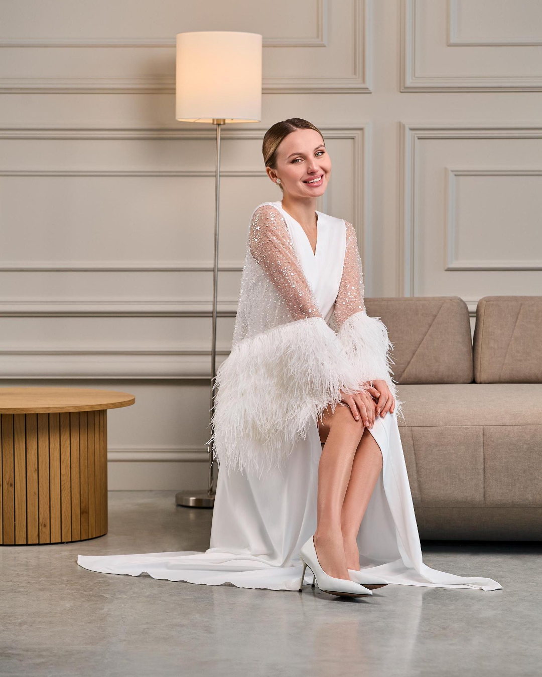 A bride poses confidently in a white satin bridal robe with sheer beaded sleeves and dramatic ostrich feather cuffs. The robe features a deep V-neckline, a waist-cinching belt, and a high slit, creating a luxurious and elegant silhouette.