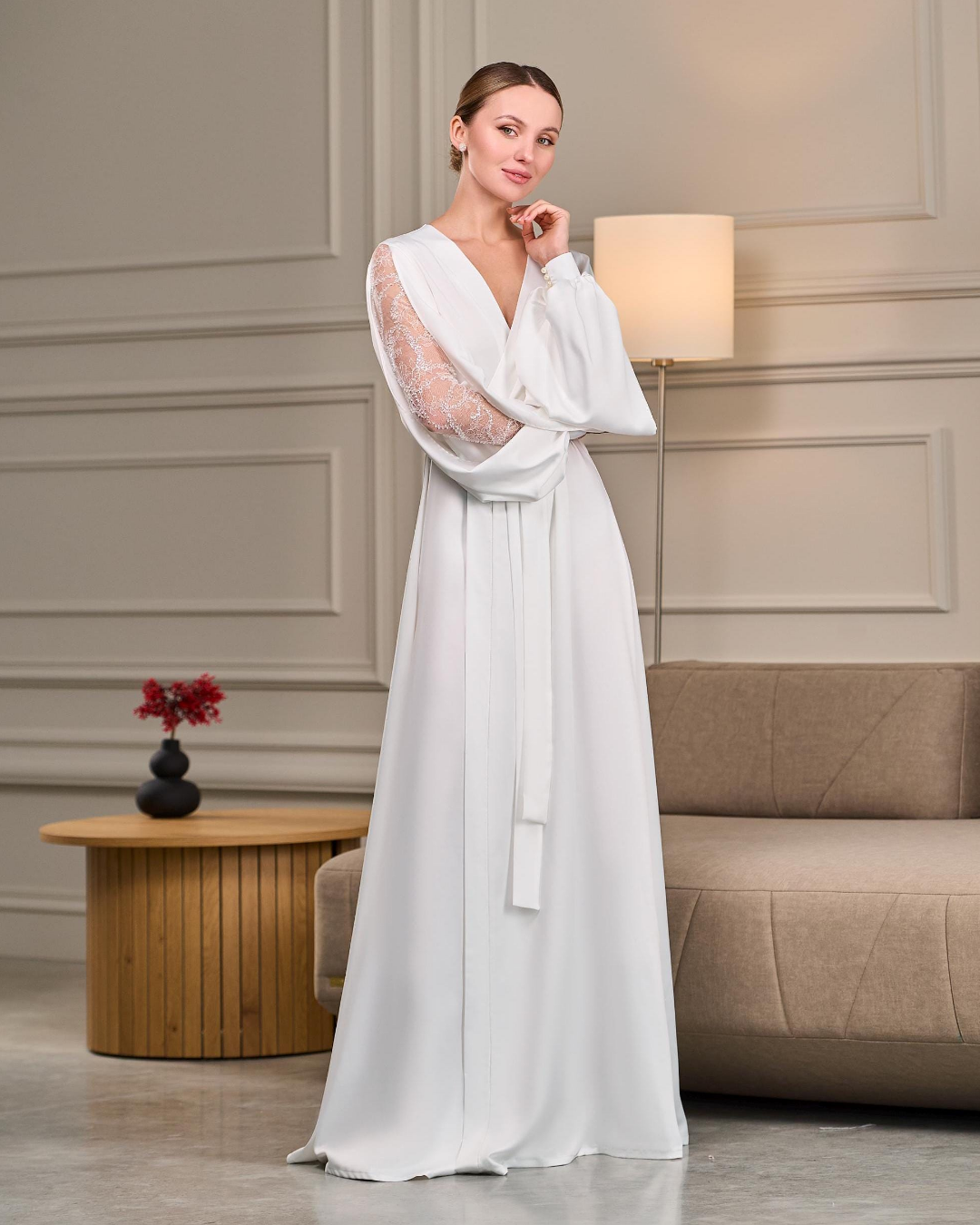 A floor-length white satin bridal robe elegantly draped on a model. The robe features sheer beaded Chantilly lace sleeves with a soft split for added movement. A silky belt cinches the waist, creating a feminine silhouette, while the flowing fabric adds a touch of effortless grace.