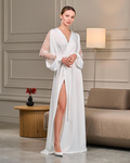 A bride gracefully poses in a luxurious white satin robe with a side slit, revealing a glimpse of her leg. The sheer lace sleeves, adorned with delicate beadwork, contrast beautifully with the smooth satin. The robe’s relaxed yet elegant fit makes it perfect for wedding morning preparations or honeymoon lounging.