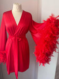 A red satin robe with long sleeves and a large feather on the side, displayed on a mannequin.