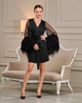 A woman wearing a black dress with feather sleeves stands in front of a white couch.
