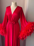 A mannequin wearing a long, red satin robe with feather accents, displayed in a room with a window.