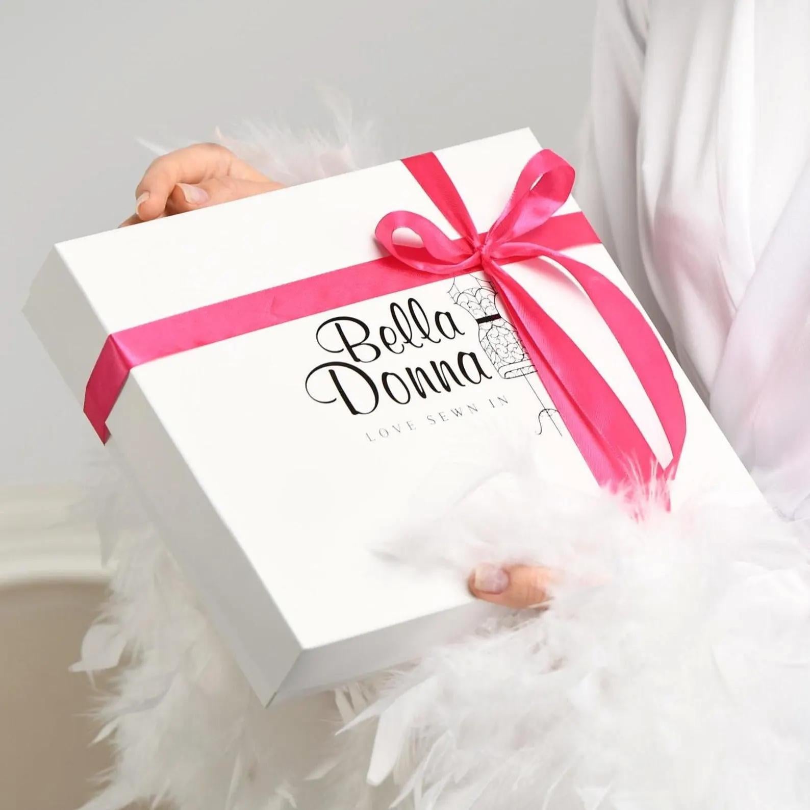 A person is holding a white gift box with a pink ribbon and the text "Bella Donna" printed on it.
