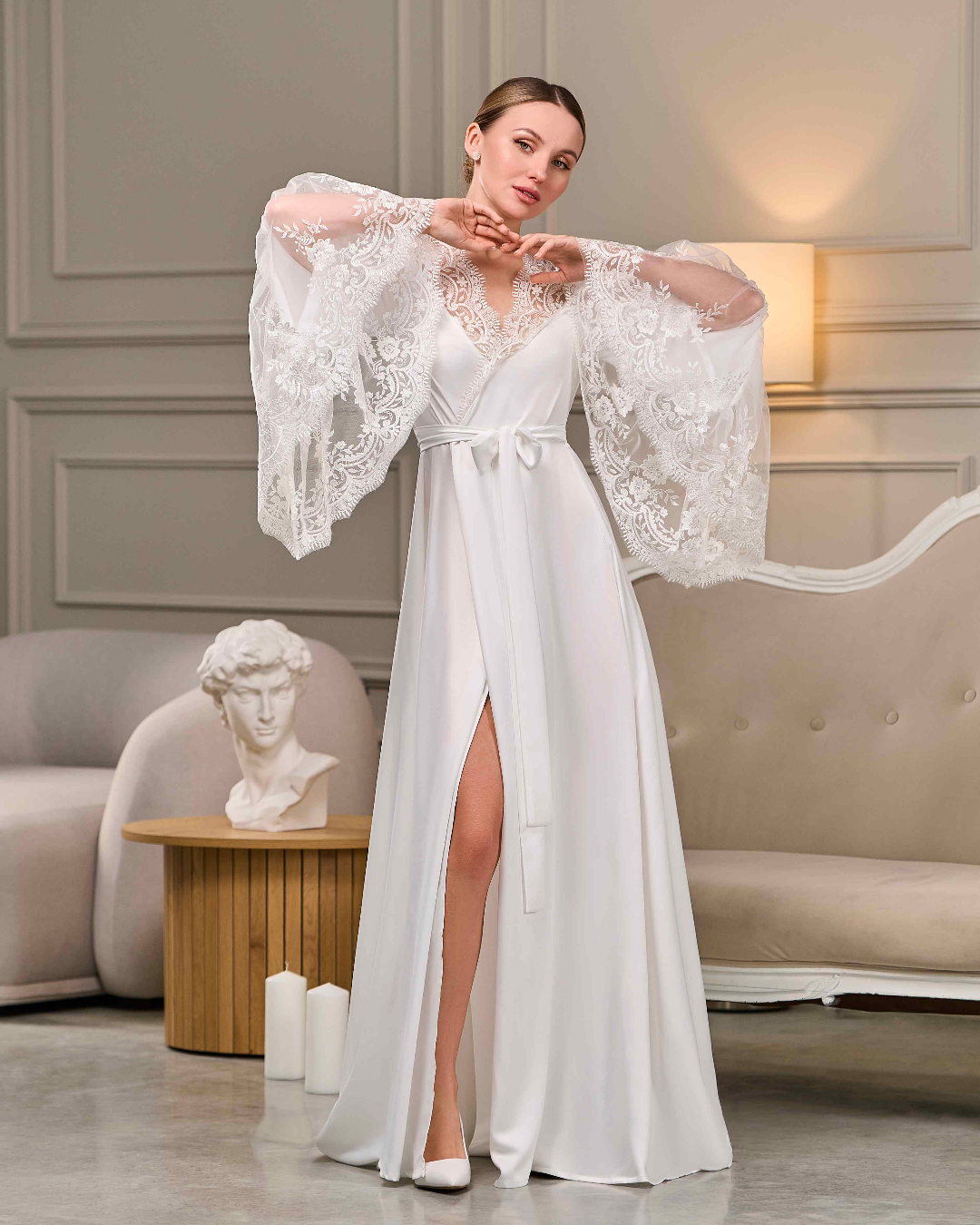 Bride in a long white robe with train and floral lace sleeves, standing in a classic elegant interior