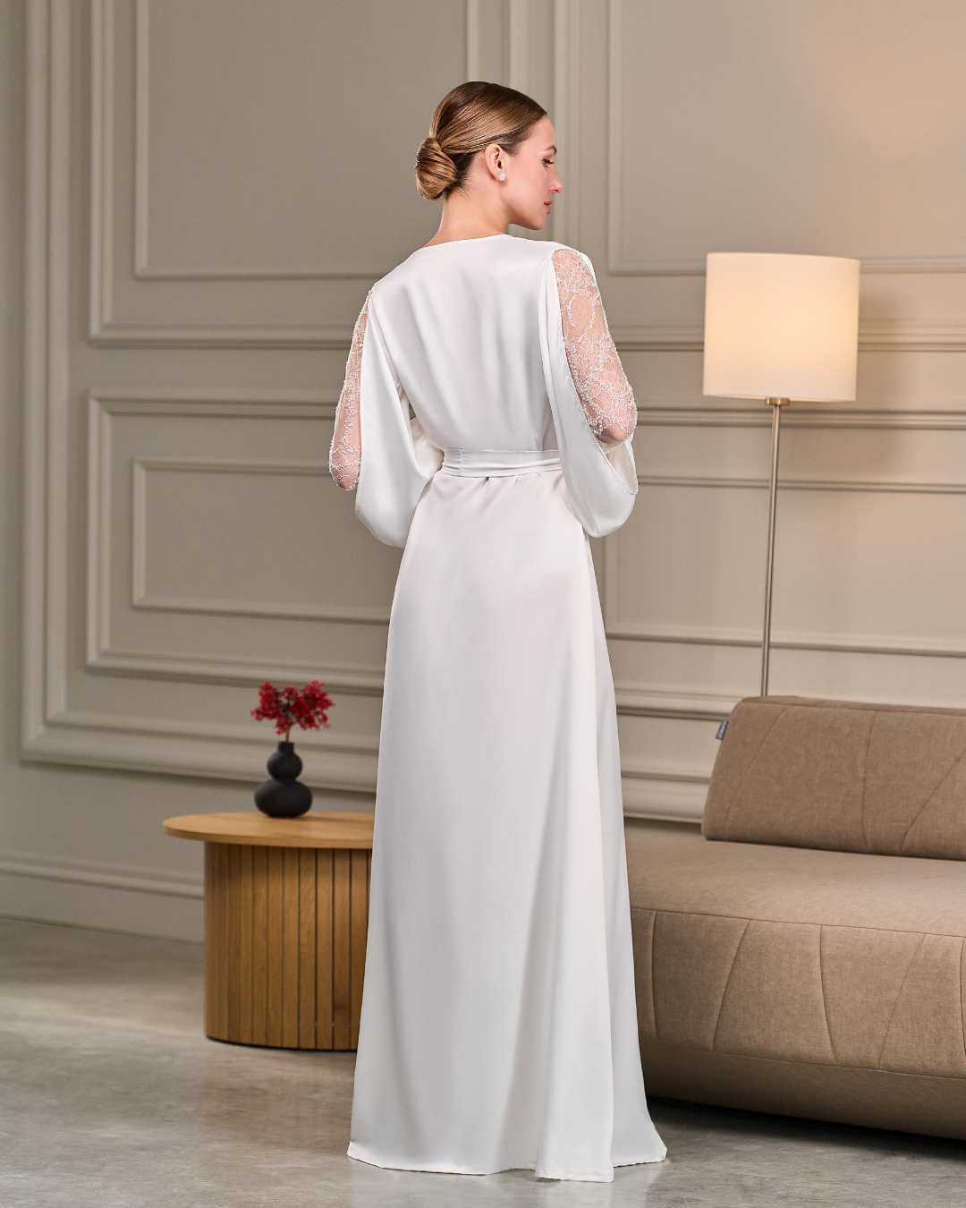 A rear view of an ethereal white satin bridal robe flowing to the floor. The sheer lace sleeves add texture and elegance, while a satin belt accentuates the waist. The soft, luxurious fabric drapes effortlessly, making it a perfect choice for brides seeking comfort and sophistication.
