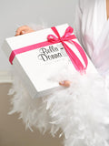A bride wearing a luxurious white satin robe with feather-trimmed sleeves holds an elegant gift box wrapped with a vibrant pink ribbon. The box features the brand name "Bella Donna" in stylish black script. The soft, delicate feathers on the robe add a touch of glamour, making this the perfect bridal gift presentation. The scene exudes sophistication and elegance, ideal for wedding mornings or special occasions.