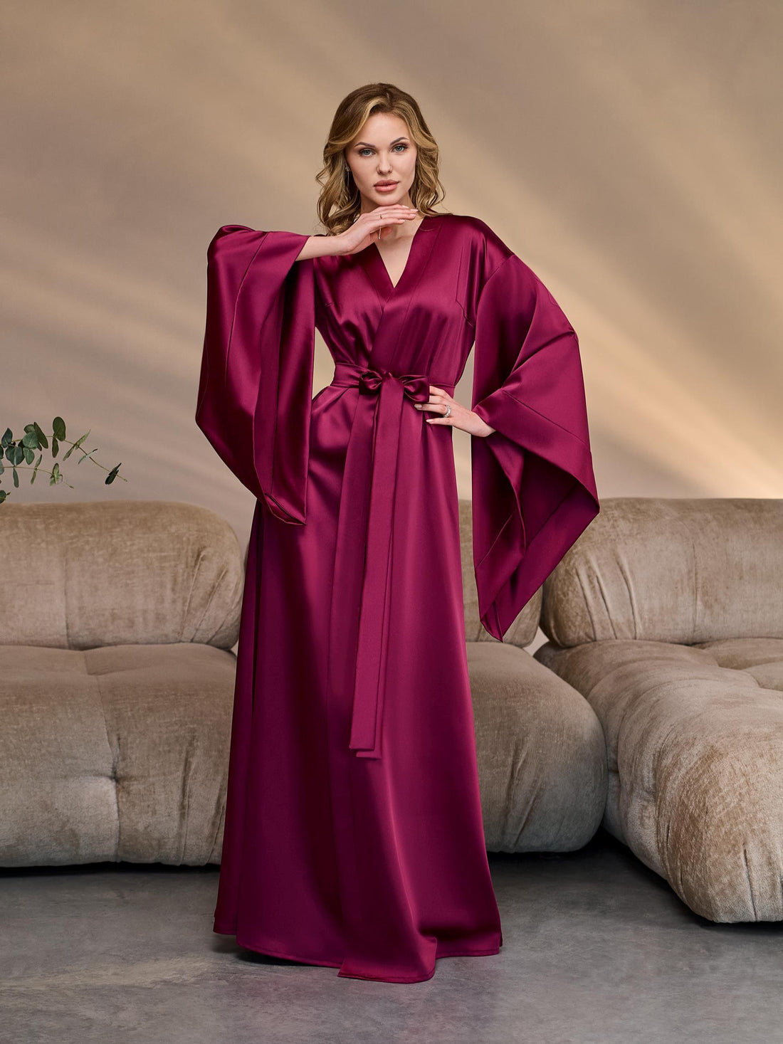 Elegant woman in a long burgundy satin kimono robe with wide dramatic sleeves, posing with raised arms to showcase the luxurious drape and high slit, perfect for bridal mornings or boudoir photography.
