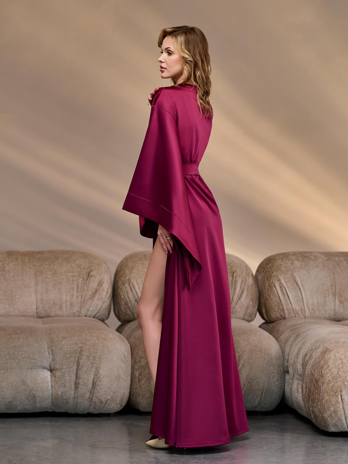 Side profile of a woman in a floor-length burgundy satin kimono robe with flowing wide sleeves and a high thigh slit, emphasizing the robe’s elegant tailoring and smooth fabric for luxury bridal styling.